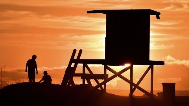 Duas pessoas sentadas na praia ao lado de um posto salva-vidas enquanto o sol se põe