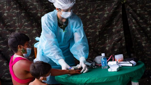 Com pano camuflado ao fundo, enfermeira prepara doses, ao lado de adulto e criança indígenas sentados - todos de máscara