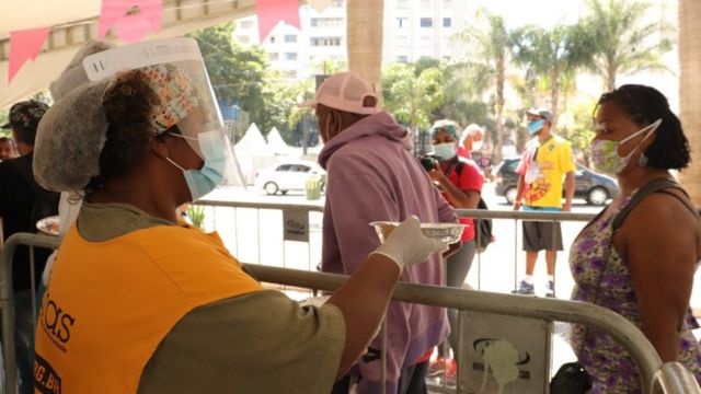 Debaixo de toldo, mulher com itens de proteção contra o coronavírus entrega uma marmita para pessoa em fila