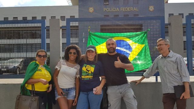 Manifestantes anti-Lula posam para foto, com bandeiras do Brasil e camisetas verde-e-amarela, em frente ao prédio da PF do Paraná