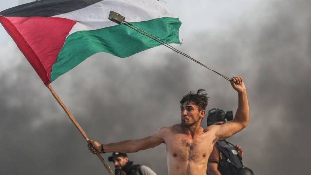 شاب يحمل العلم الفلسطيني بيد وفي الأخرى مقلاع يرمي به الجنود الإسرائيليين