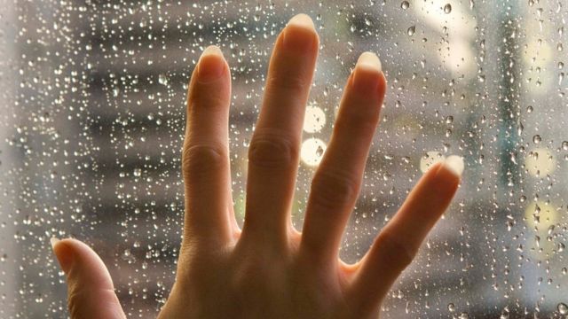 Una persona con la mano sobre una ventana, mientras afuera llueve