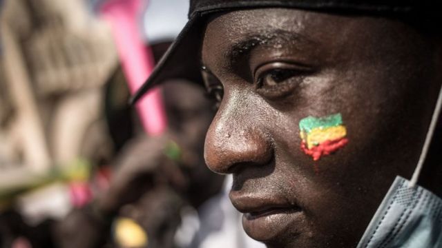 Un manifestant observe la foule avec un drapeau malien peint sur la joue, lors d'une manifestation de masse à Bamako, le 14 janvier 2022, pour protester contre les sanctions imposées au Mali et à la junte par la Communauté économique des États d'Afrique de l'Ouest (CEDEAO)