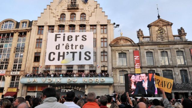 Prefeitura no norte da França com uma multidão e um grande banner com os dizeres 'Orgulho de ser dos Ch'tis'