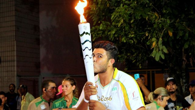 Ygor Marcel da Cruz Santos segura com as duas mãos e beija a tocha olímpica, vestido com o uniforme branco e amarelo utilizado pelos condutores nas Olimpíadas de 2016. Ao fundo, há outras pessoas e a folhagem de uma árvore
