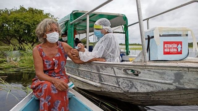 Profissional da saúde aplica vacina no braço de mulher, que está num barco