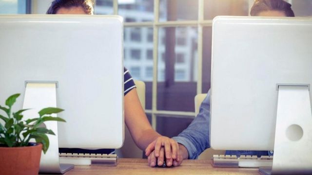 Un hombre y una mujer cuyos rostros no se ven, por estar detrás de unas pantallas de computadora, pero que se toman de la mano