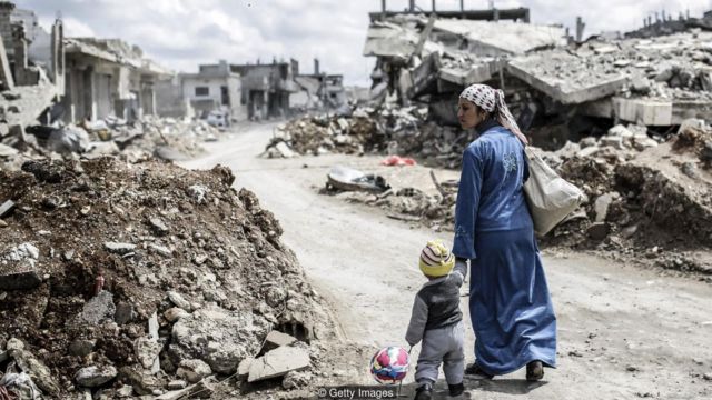 Mulher caminha com criança em meio a ruínas de cidade na Síria após conflito entre combatentes