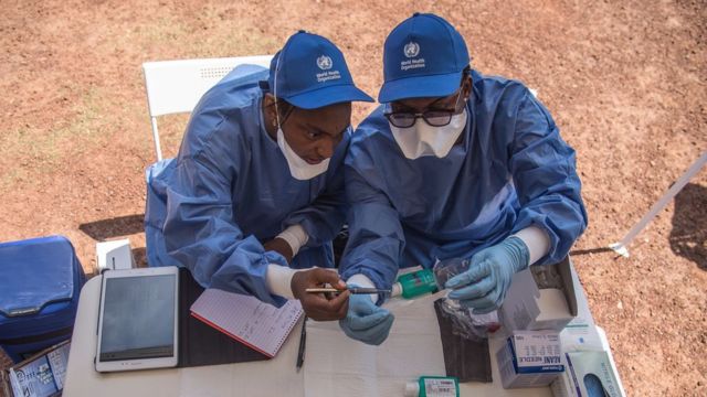 Duas enfermeiras da Organização Mundial de Saúde, com uniformes azul-claro, boné e luvas, se preparam para administrar vacinas