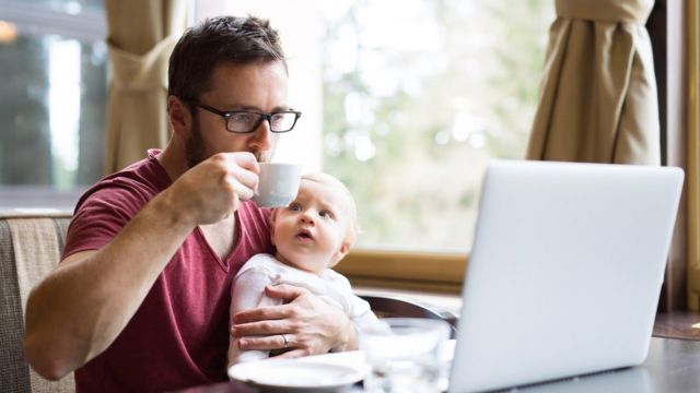 Hombre tomando un café, con un bebé en brazos frente a una computadora.