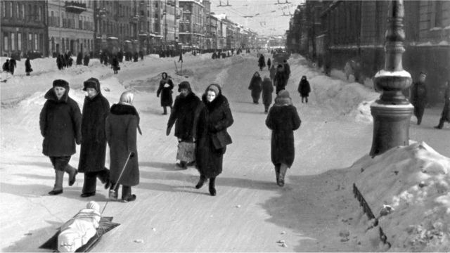 Des habitants marchent dans les rues de Leningrad assiégée. L'un d'eux tire le corps d'un parent mort sur un traîneau.