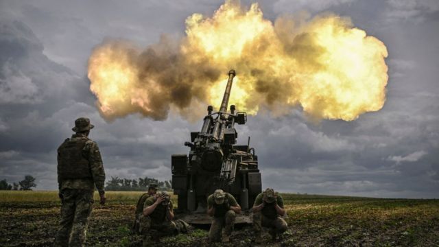 Fotografia colorida mostra homens com uniforme militar operando um canhão militar que solta fogo em um campo verde