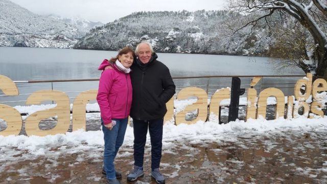 O casal de idosos Stella e Norberto posa para a foto em frente a um um lago e montanhas baixas cobertas de neve.