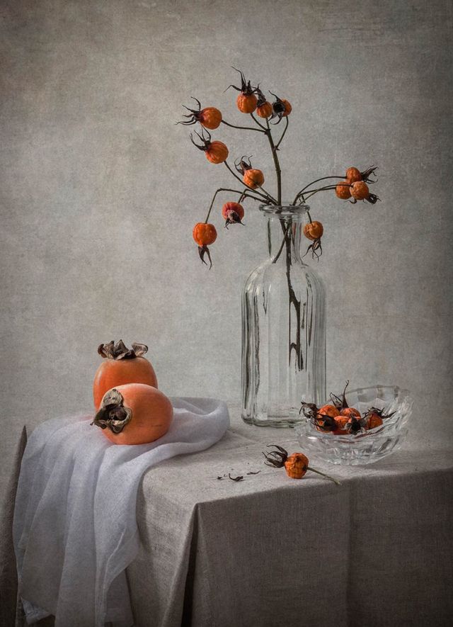 Rose hips in a glass vase with ripe persimmon fruits on table with a grey tablecloth