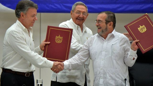 Juan Manuel Santos estrecha la mano de Timochenko mientras Raúl Castro sonríe.