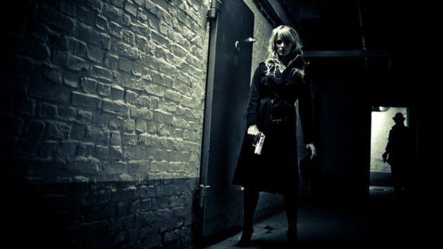 A woman spying with a gun in a hallway observed by a man in the shadow of a door