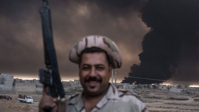 An Iraqi soldier holds a rifle as smoke billows from the Qayyarah area,
