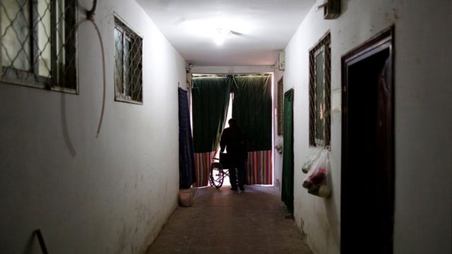 Hombre empuja una silla de ruedas a través de unas cortinas oscuras, al final de un pasillo.
