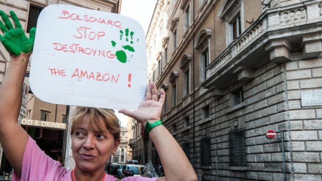 Mulher segura cartaz de protesto em inglês, onde se lê: Bolsonaro, pare de destruir a Amazônia