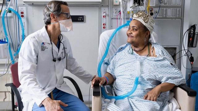 Médico sentado ao lado de paciente em maca, ambos sorrindo e conversando