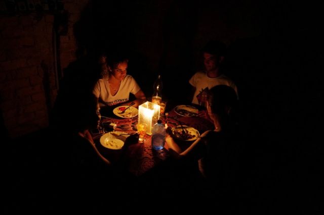 Tres personas de una familia sentdas a la mesa, todo está a oscuras y se alumbran con velas.
