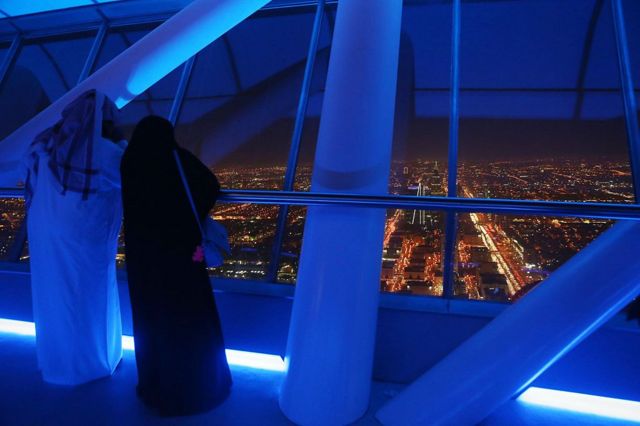 Dos personas observan una panorámica nocturna de Riad desde lo alto de un rascacielos. 