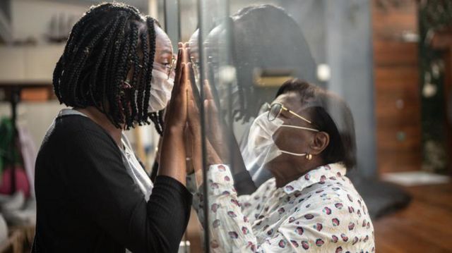 Duas mulheres de m&aacute;scara interagem atrav&eacute;s de um vidro