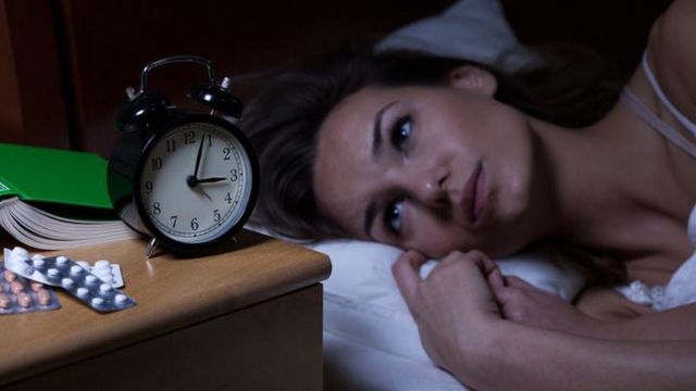 Mulher deitada na cama olhando para relógio e cartelas de comprimidos