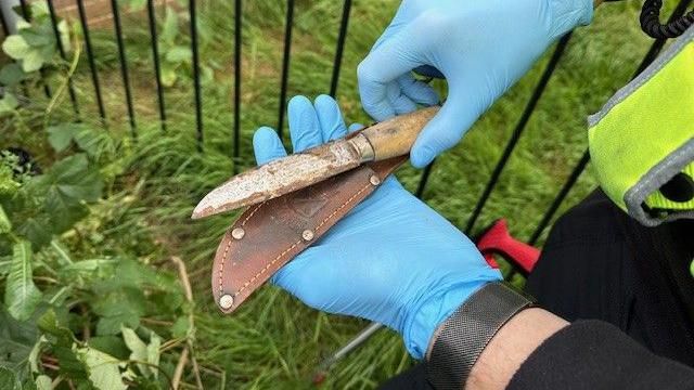 A rusted knife and sheath being held in gloved hands