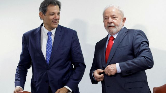 Haddad e Lula lado a lado, em pé, em ambiente interno. Haddad olha para Lula sorrindo