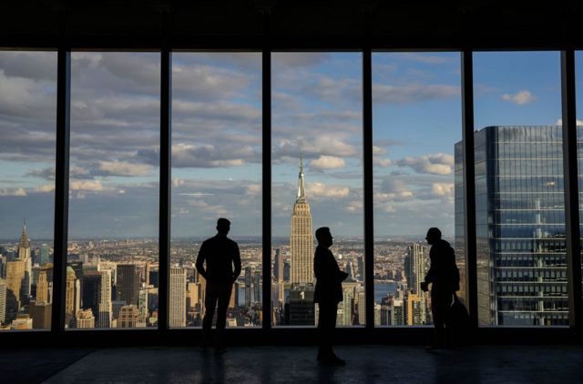 Silhuetas de três pessoas contra a janela em um escritório em Nova York. Ao fundo vê-se a cidade e o Empire State Building
