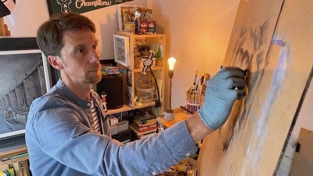 Matt Sayers, dressed in a blue short and wearing a latex glove as he draws with charcoal at an easel in his workshop by a bare lightbulb