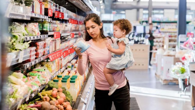 Uma mãe segurando a filha e olhando os preços dos produtos em um supermercado