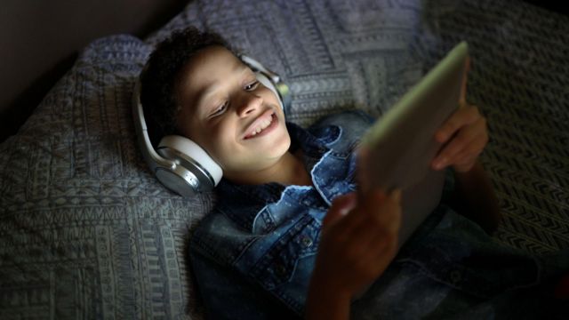 Criança sorrindo enquanto assiste algo na cama, com table e fone de ouvido