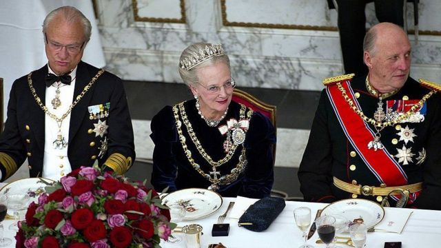 Três idosos brancos com roupas oficiais da realeza em frente a uma mesa de jantar