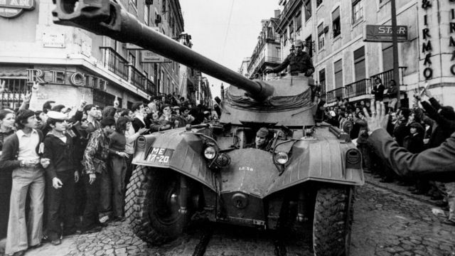 Foto em preto e branco mostra dezenas de pessoas na rua olhando para tanque, que passa no meio da via