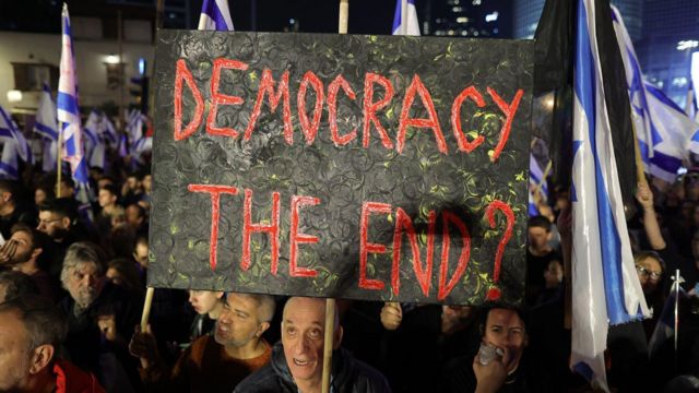 Foto noturna mostra várias pessoas na rua com bandeiras de Israel; homem aparece com cartaz dizendo em inglês: 'Democracia, o fim?'