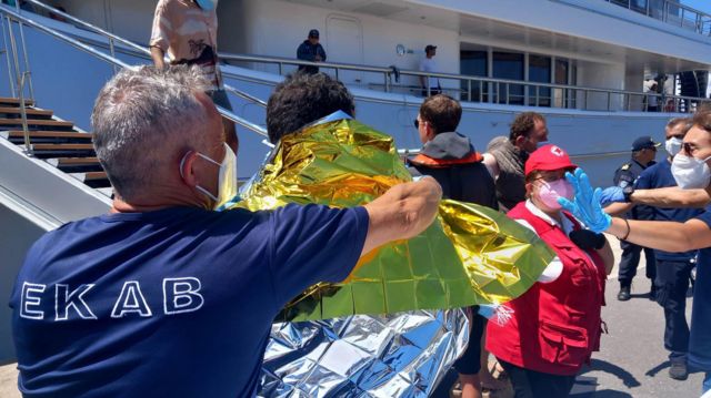 Agente com mscara coloca manta trmica sobre homem, que aparece de costas e perto de grande barco