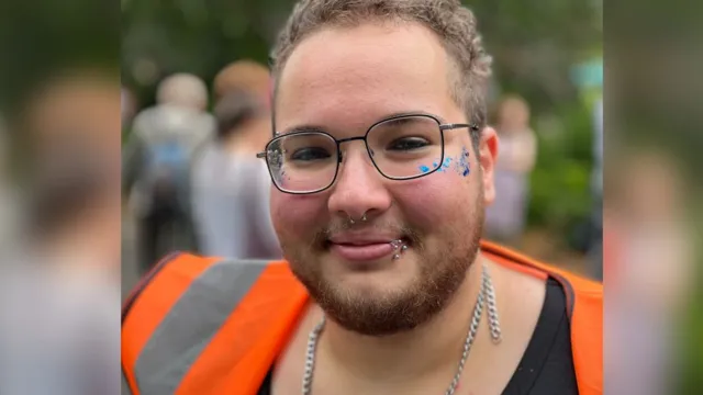 Nathan Maradei, wearing a hi-vis vest over a black shirt and glasses, smiles with blue glitter on his cheeks.