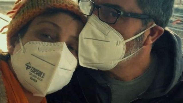 Claudia e o Marido com os rostos próximos, de máscara, olhando para a câmera