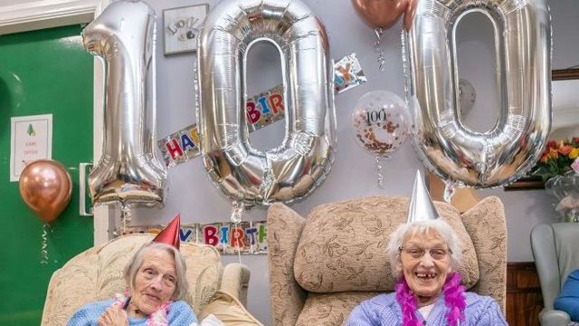 Duas senhoras sentadas sorrindo em frente a balões em formato de 100