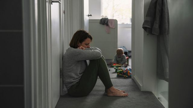 Mãe sentada no corredor, com postura de cansaço, e filho bebê brincando atrás