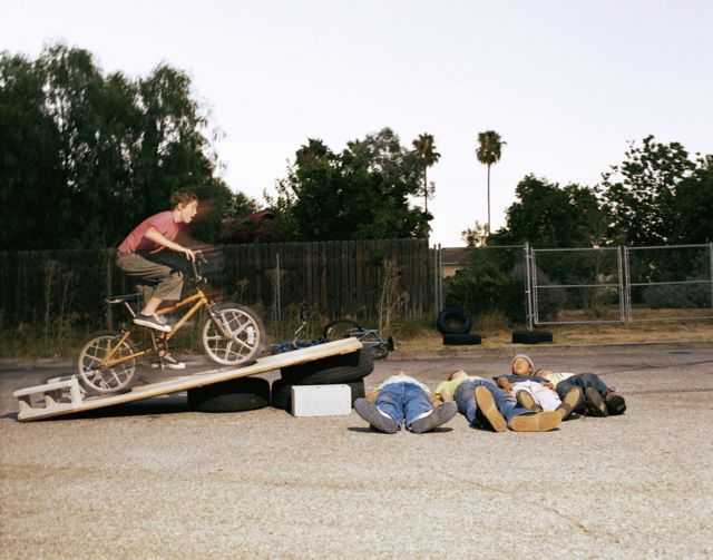 Adolescente branco em rampa de bicicleta; embaixo da rampa estão deitados outros 4 adolescentes
