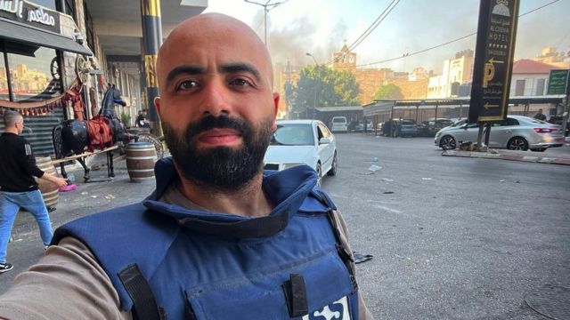 Alaa Daraghme com colete a prova de balas em rua, fazendo selfie, com fumaça atrás