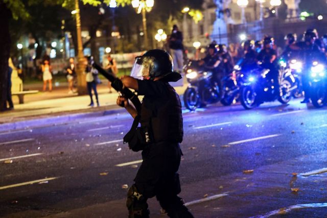 Policial com arma de bala de borracha e motos de policiais atrás