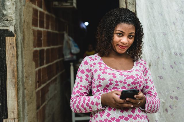 Mulher jovem de baixa renda segurando um celular e sorrindo timidamente para a câmera