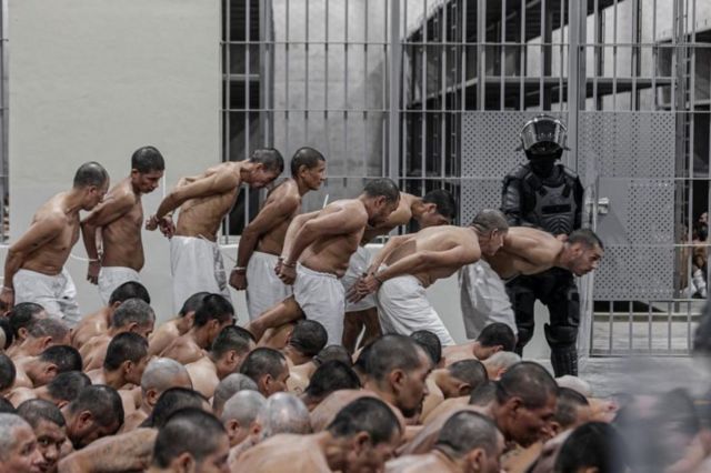 Presos andando agachados em fila indiana enquanto so observados por policial