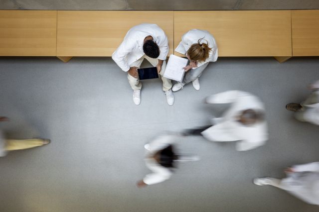 Cientistas de jaleco branco caminhando em corredor e outros sentados, conversando e olhando para caderno