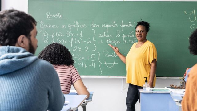 Foto mostra professora negra de pé dando aula para uma classe de alunos jovens e diversos de costas