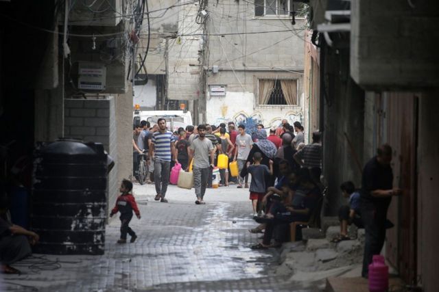 Crianças e adultos em rua, alguns deles carregando galões de água